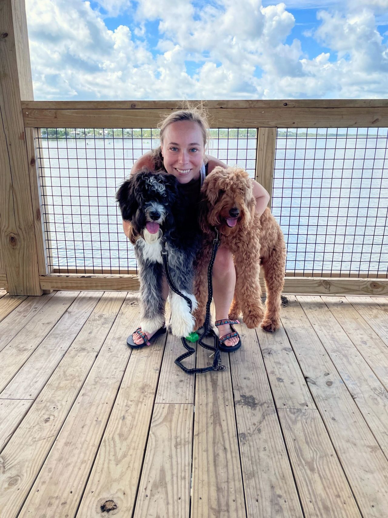 A woman with light-brown hair crouches on a waterfront deck hugging Otto and Remi, with a blue sky and water in the background
