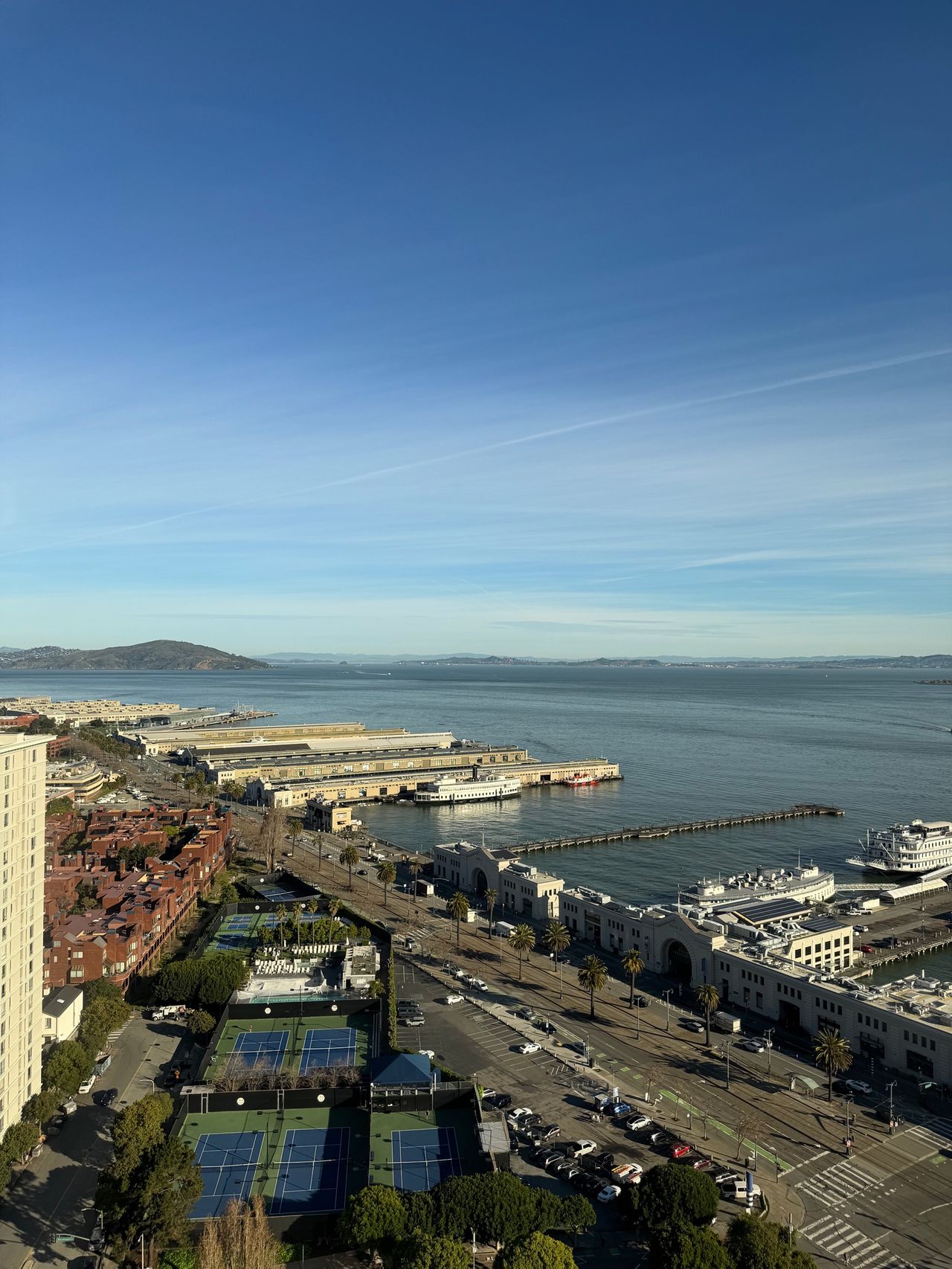 An elevated view of San Francisco Bay from a high floor at 4 Embarcadero Center, looking past long piers, tennis courts, and a residential block toward distant headlands.