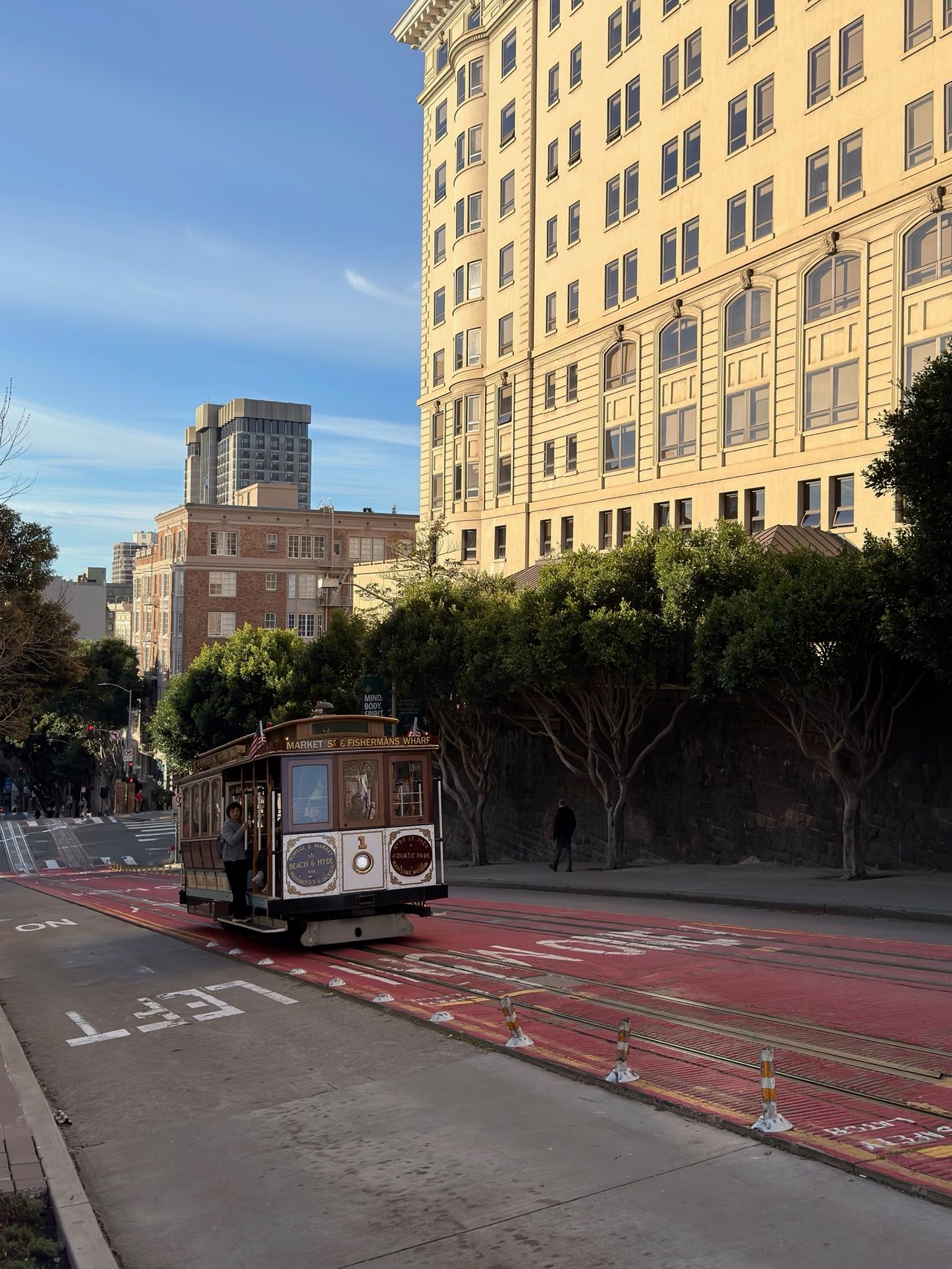 A vintage cable car crests a hill on California Street, framed by a tall beige hotel and a brick-painted right-of-way down the middle of the road.