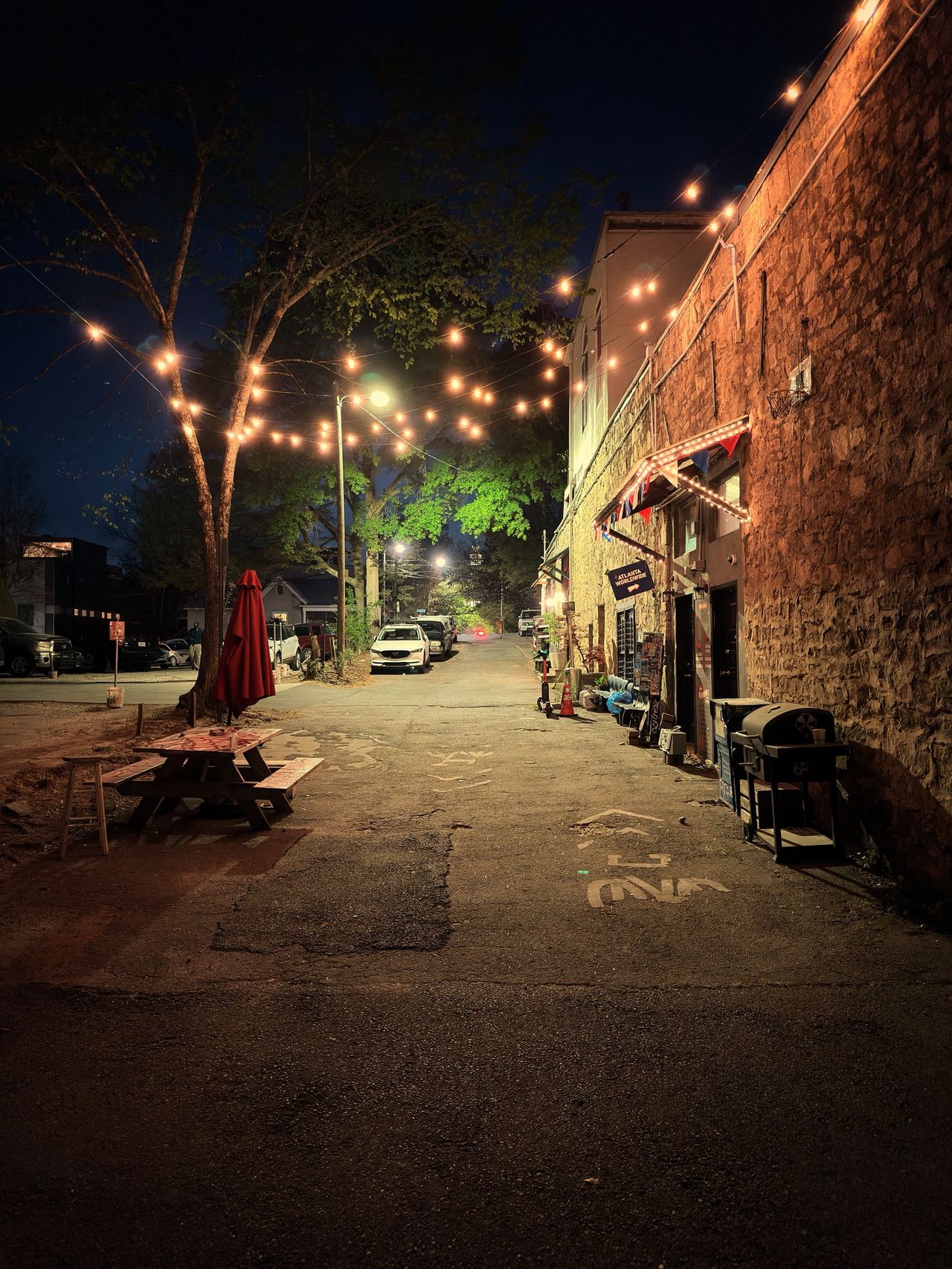 A nighttime alley with picnic tables under a tree strung with festoon lights, a brick building on the right with festive bunting, and parked cars in the distance.