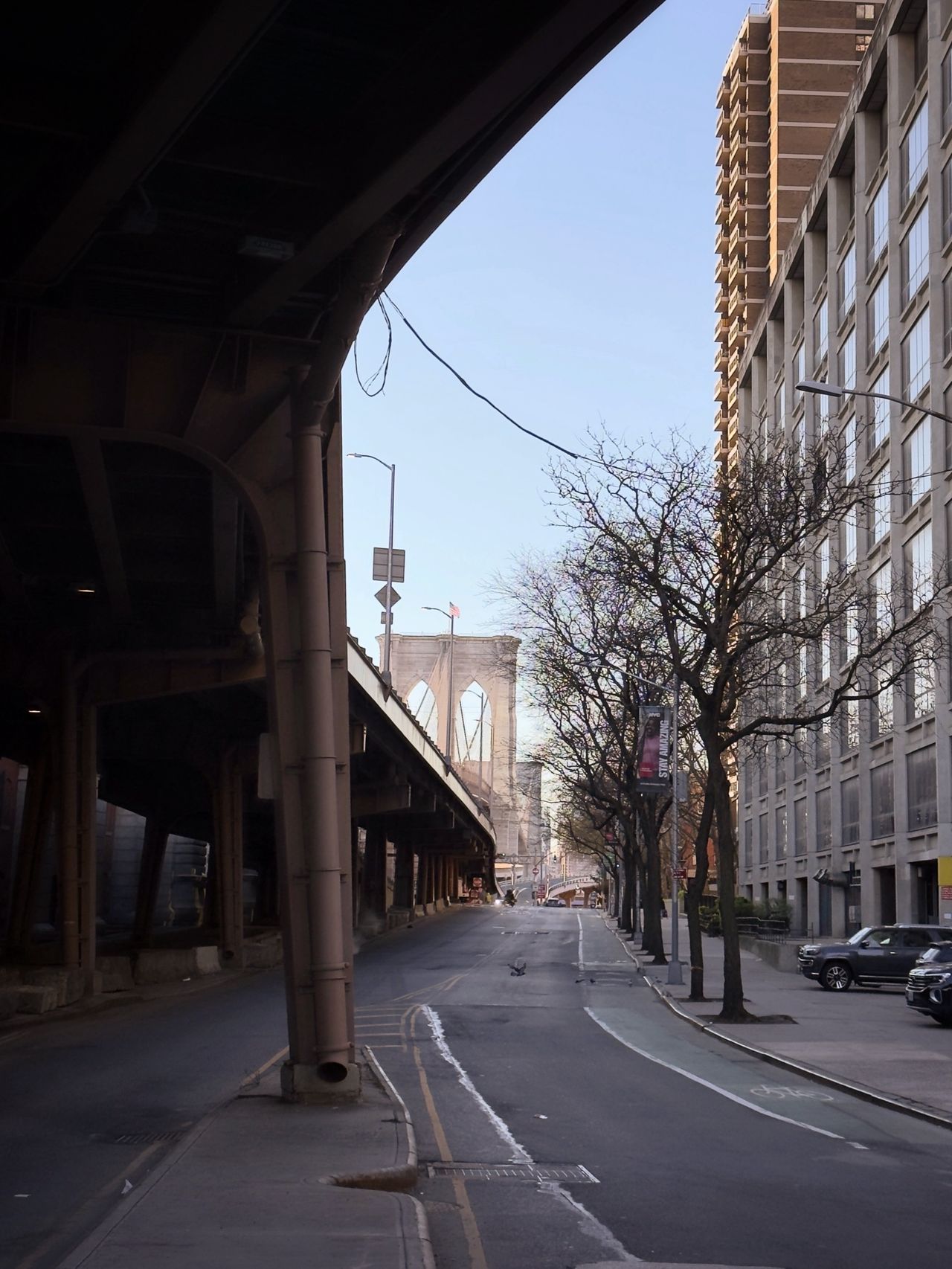 Looking out from beneath an elevated highway toward a stone tower of the Brooklyn Bridge, framed by an iron support column.