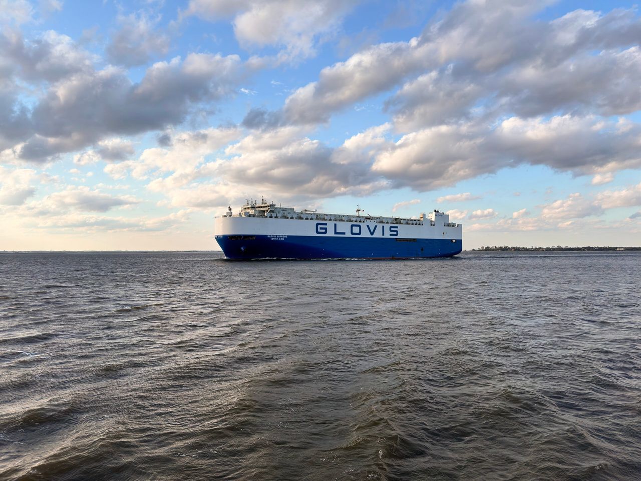 A large car carrying cargo ship moves through the channel just before sunset. It is gray and blue two-tone with GLOVIS written in large letters across the side.