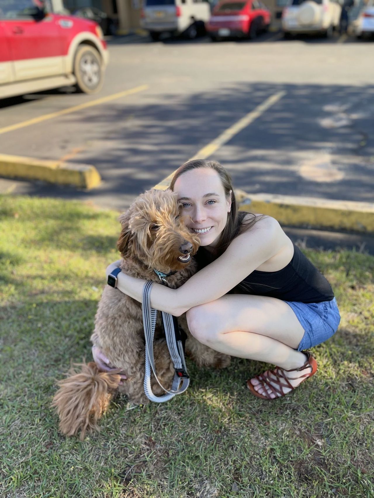 Holly, in a black sleeveless top and shorts, shares a heartfelt embrace with Remi, her brown furry friend, on a sunlit patch of grass. Both of them smile.