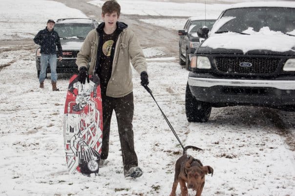 Luke holds a boogie board while his dog pulls his leash in the snow.