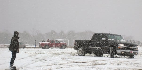 Someone on waits on a snowboard in the foregronud a black and red truck in the distance.