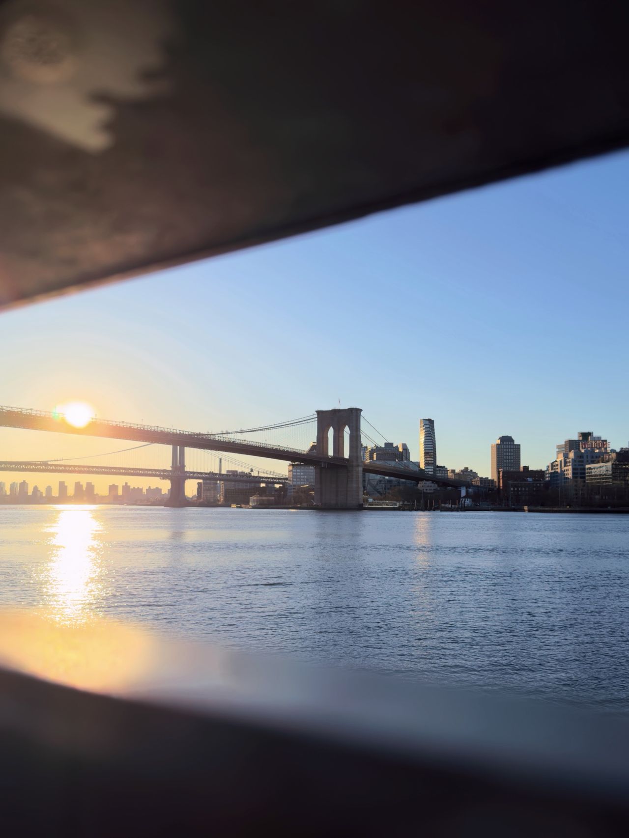 Brooklyn Bridge silhouetted against a rising sun, framed between dark ferry railings over the East River