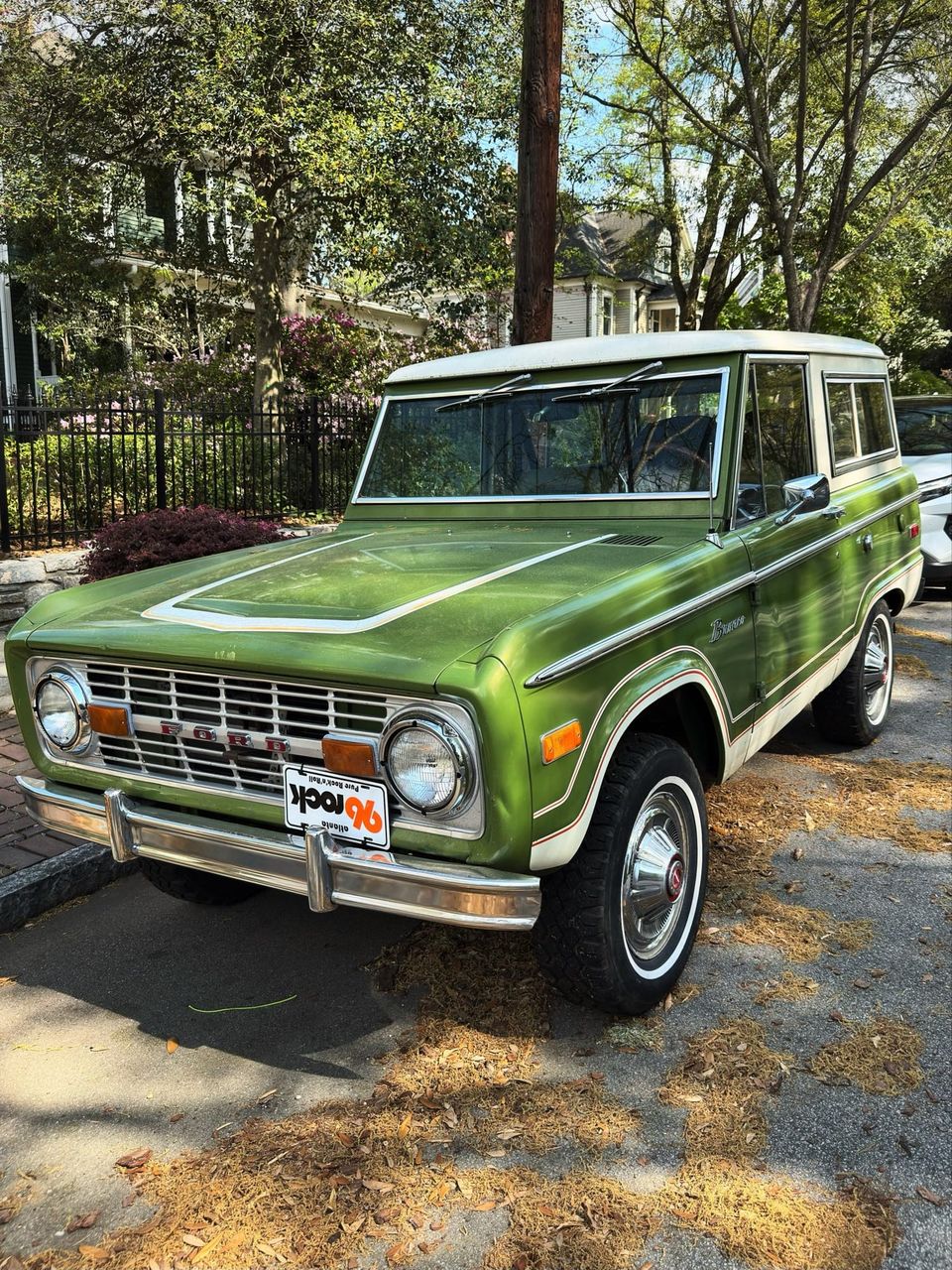 Green and white vintage Ford Bronco parked on a tree-lined street with a 96 Rock plate on the front bumper