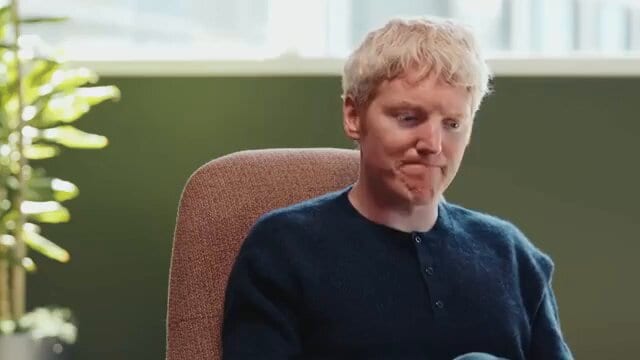A blond man in a dark sweater sits in a chair, looking down with a slight smile