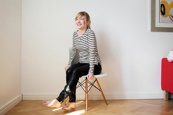 A smiling woman with short blonde hair in a striped top and leather pants sitting barefoot on a white stool