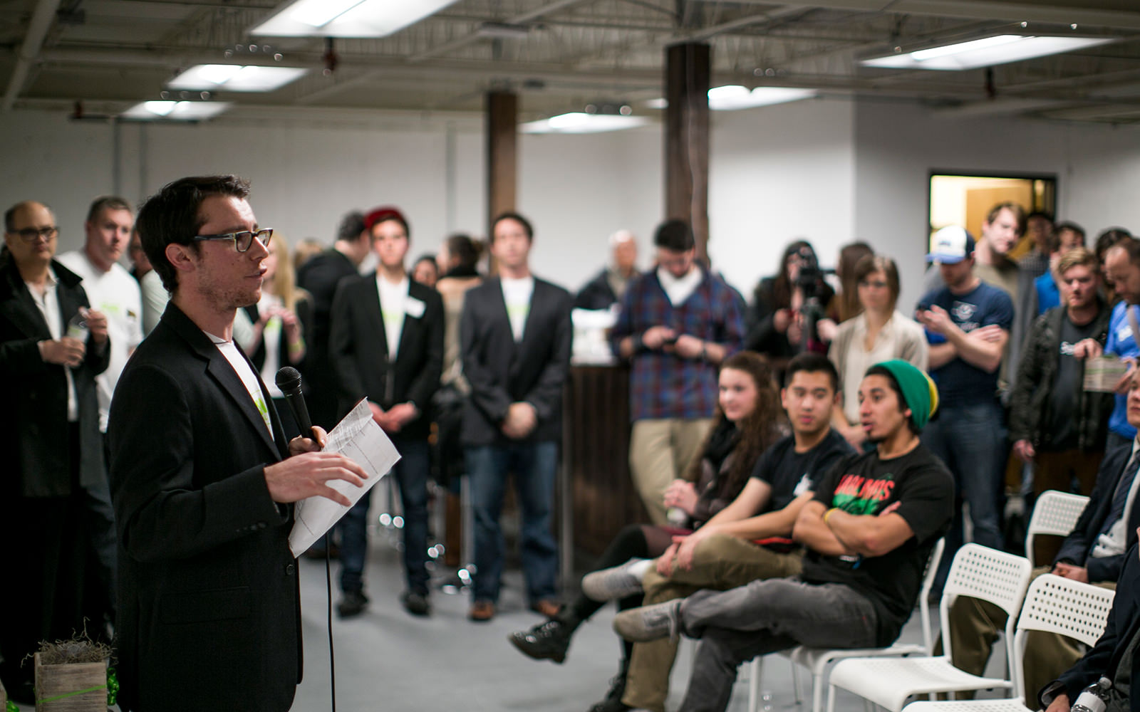 Sam Solomon speaking into a microphone and holding notes at an indoor event with a crowd of attendees in the background
